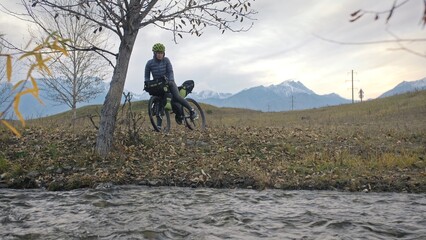 The woman travel on mixed terrain cycle touring with bikepacking. The traveler journey with bicycle bags. Sportswear in green black colors. The trip in magical autumn forest, river stream.