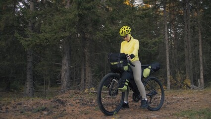 Naklejka premium The woman travel on mixed terrain cycle touring with bike bikepacking outdoor. The traveler journey with bicycle bags. Stylish bikepacking, bike, sportswear in green black colors. Magic forest park.