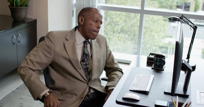 A serious corporate black man rubbing his forehead suddenly angrily slams the desk in frustration while working at a desktop computer near a large office window. 35mm Medium Closeup Tripod Shot 4K.