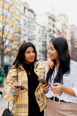 Two Latin women looking at each other and holding their mobile phones in the street. Hispanic sister having a good time together sightseeing.
