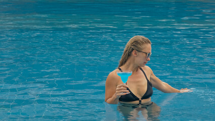 Middle shot pretty woman drinking blue cocktail alcohol liquor in swimming pool at hotel. Portrait of sexy girl in pool outdoor. Beautiful caucasian women with blond hair, black swimsuit, sunglasses.