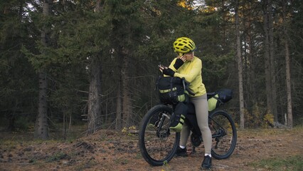 Naklejka premium The woman travel on mixed terrain cycle touring with bike bikepacking outdoor. The traveler journey with bicycle bags. Sportswear in green black colors. Magic forest park. Make a selfie smartphone.