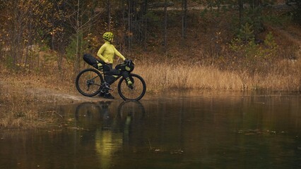 The woman travel on mixed terrain cycle touring with bikepacking. The traveler journey with bicycle bags. Sportswear in green black colors. The trip in magical autumn forest, river stream.