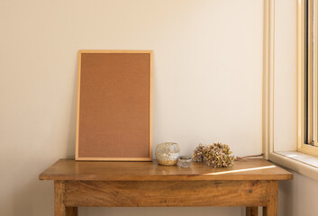 Cork board on oak sidetable with glassware and dried hydrangeas against beige wall next to window (selective focus)