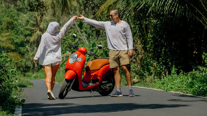 Two caucasian tourist woman man dance near scooter. Love couple on red motorbike in white clothes, sunglasses on forest road trail trip. Dancing road. Motorcycle rent. Asia Thailand ride tourism.