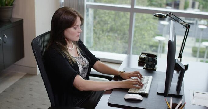 An angry and serious young woman with long brown hair works at a desktop office computer, becomes impatient, deeply sighs, and leans forward to lean on hand bored. 35mm Medium Tripod Shot 4K.