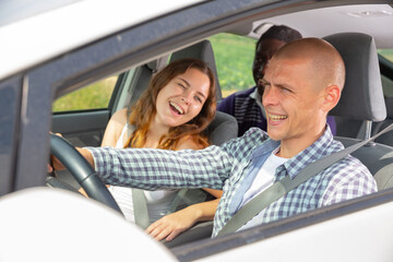 Cheerful young man driving car with laughing friends in passenger seats. Friendly road trip and travel concept