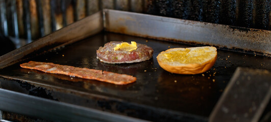 Closeup shot of delicious fresh baked burger bun with butter, minced cooked patty beef meat with margarine and pork bacon grilled on frying pan stove at dirty unsanitary local restaurant counter