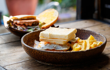 Closeup shot delicious tasty yummy homemade breakfast fried egg, pork bacon and cheese sandwich cutting edge bread with straight cut fresh frying french fries appetizer in wooden bowl on wood table