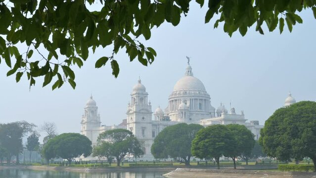4K stock video of Victoria Memorial, a large marble building in Central Kolkata, It is one of the famous monuments of Kolkata, West Bengal, India. Tree leaves hanging from above in the frame.
