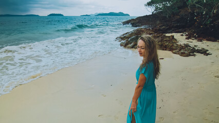 Alone attractive woman walk in beach, near a rock reef hill in stormy morning rain cloudy sea. Girl walk in turquoise swimsuit. Concept resort outdoor relax, vacation, loneliness. Dark dramatic view
