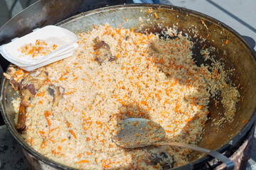 A mountain of delicious rice pilaf with large pieces of meat in a cauldron on a fire in the open air. Street food at public events.