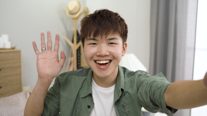 Korean boy is waving hi and touching his face with a funny expression while talking with friends on a video call in a bright home interior. © PRPicturesProduction
