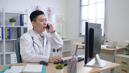asian male physician working in front of the computer is talking in surprise and leaving his office right away as soon as he receives an emergency call.