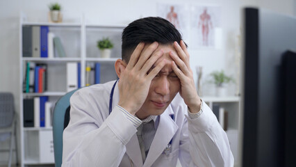 asian male physician under pressure at work is burying his face with hands and breathing a deep sigh while looking at the computer monitor with anxiety.