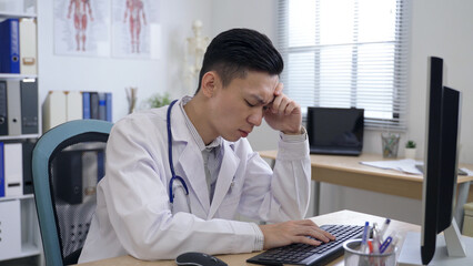 asian doctor experiencing stress at work is propping head and touching his face while dealing with a challenging problem on computer at office desk