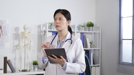 concentrated asian physician is glancing at the distance and pondering with a serious look while doing a research on her tablet computer in her office