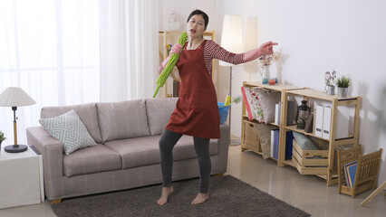 cheerful young wife wearing an apron is singing into the feather boom and shaking her body during housecleaning at home living room.