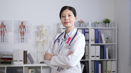 breast cancer awareness concept physician looking at the camera and crossing her arms is wearing a medical uniform with a pink ribbon in the office.