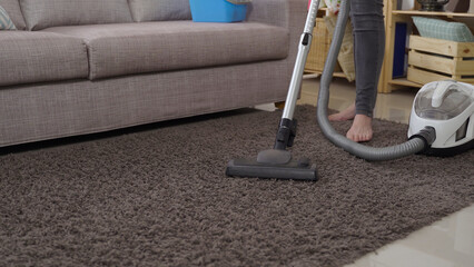 close up view vacuum cleaner head moving back and forth on the gray rug by the sofa with a woman standing barefoot in the background.
