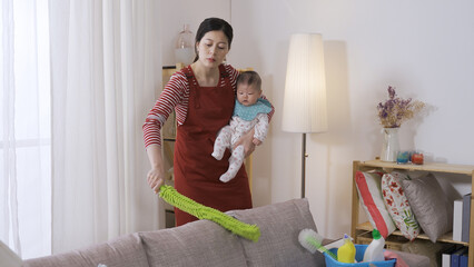 Chinese full time mother is talking to her young child in arm while beating and sweeping the sofa with a duster broom in the living room.