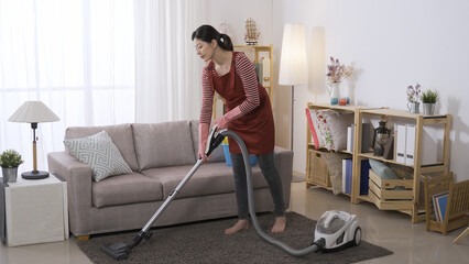 full length asian woman is slowly vacuuming the rug while doing housecleaning in a bright living...