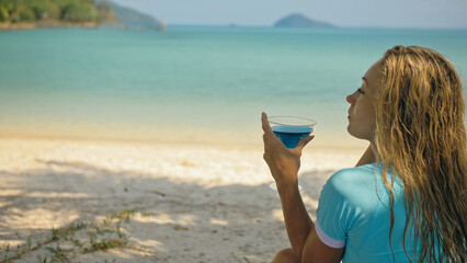 The woman is holding a glass of blue curacao cocktail, on beach azure blue sea of a tropical island. Blurred beautiful seaside background. Celebrate summer holiday concept of leisure, travel, relax.