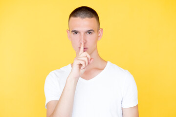 Keep secret. Silent man. Mute expression. Serious hipster guy in casual white t-shirt holding shush finger on mouth isolated yellow.