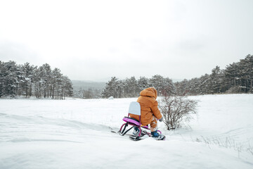 Little toddler child in winter outfit sits on sledge in snowy park