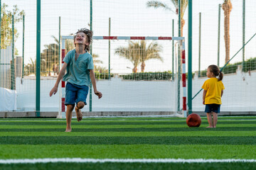 Happy boy running along green lawn and laughing during playing football and little girl standing not far 