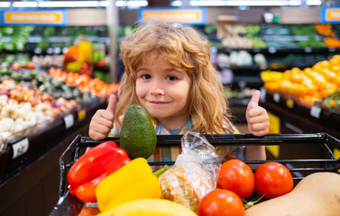 Child in supermarket buying fruit. Kid grocery shopping. Kid with cart choosing fresh vegetables in local store. Boy having fun while choosing food in the supermarket.