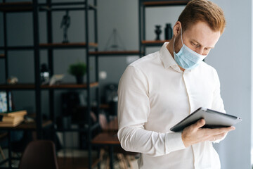 Portrait of focsued young business man wearing protective face mask using digital tablet standing in modern office room, looking on screen. Confident employee using smartphone planning work time.