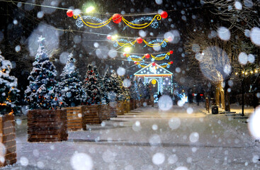City street during snowfall at winter night. Beautiful illumination and decoration on street. New Year Christmas holidays celebration. Lanterns garlands on trees.
