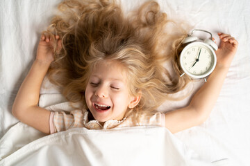 happy little blonde girl in a white bed with an alarm clock, good morning