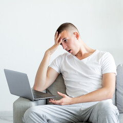 Clueless situation. Confused man. Online conference. Displeased shocked casual guy sitting sofa looking laptop facepalm hand on head in light room interior.