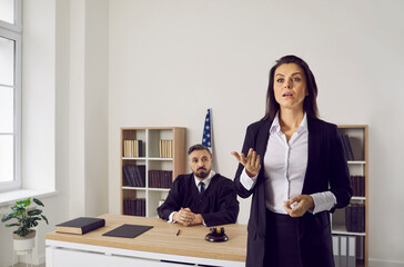 Attorney pleading in court. Professional female lawyer appears before jury and provides evidence during trial. Serious lawyer speaks against the background of a male judge sitting at a table.