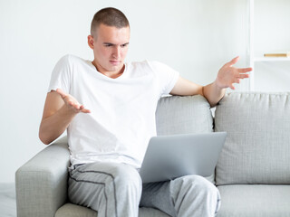Uncertain situation. Hesitating man. Online meeting. Confused casual guy sitting sofa looking laptop with shrugging hands in light room interior.