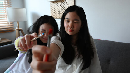 Two playful asian girls holding paintbrushes and smiling at camera.