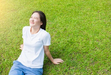 Asian girl in white shirt Come out to relax and enjoy the warm sunlight in the evening On a happy rest day from work Outside sitting on the green lawn, looking up at the light and smiling in the park