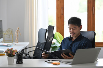 Asian male entrepreneur sitting in modern workplace and checking financial document.