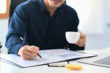 Young businessman holding cup of coffee and checking financial reports at office desk.