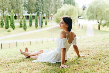 Young pretty woman with cute smile resting, relaxing sitting on green grass in park. Side view of brunette lady in white summer dress enjoying chilling and looking away, outdoors