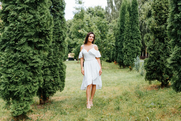 Full length portrait of pretty young brunette woman in white dress with cute smile looking away in park. Elegant lady model with handbag standing outdoors on summer day. Fashion and beauty