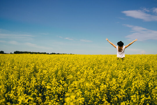 the woman in the hat enjoying nature in the middle of field full of yellow flowers. Yellow field of flowering rapeseed with cloudy blue sky - brassica napus - plant for green energy, medicine.
