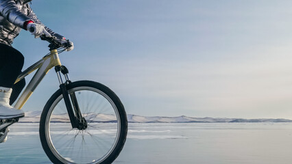 Fototapeta premium Woman is riding bicycle on the ice. Girl is dressed in a silvery down jacket, cycling backpack and helmet. Ice of the frozen Lake Baikal. Tires on bike are covered with spikes. Traveler is ride cycle.