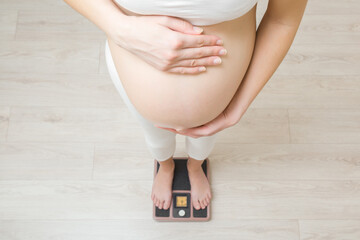 Young adult pregnant woman standing on weight scales. Hands touching naked big belly. Care about body health in pregnancy time. Closeup. Top down view. - obrazy, fototapety, plakaty