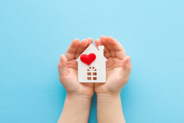 Baby opened palms holding white house with bright red heart on light blue table background. Pastel...