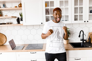 Joyful male freelancer having successful video meeting. Smiling african american male using yellow laptop, staying in kitchen at home, distant work concept,excited with good news and business success.