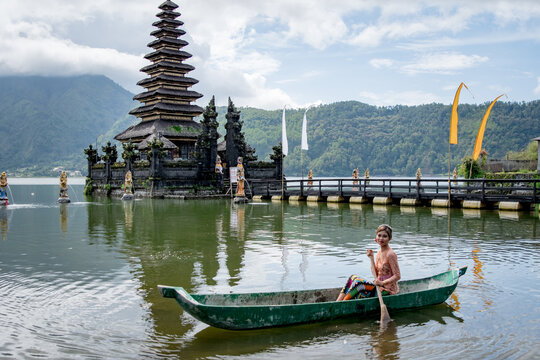 Young girl paddling on a wooden boat on a lake near a Bali temple.