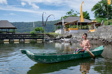 Young girl paddling on a wooden canoe in traditional clothes, and Hindu temple surrounded by body...
